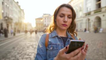 ung kvinna bär denim jacka gående ner ett gammal gata använder sig av smartphone på solnedgång. kommunikation, social nätverk, uppkopplad handla begrepp. långsam rörelse video