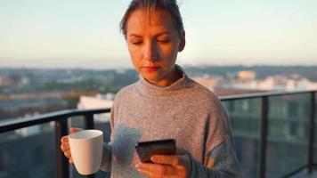 Woman starts her day with a cup of tea or coffee and checking emails in her smartphone on the balcony at dawn, slow motion. Modern urban lifestyle video