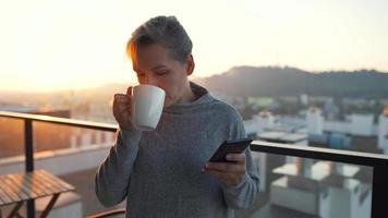 mujer empieza su día con un taza de té o café y comprobación correos electrónicos en su teléfono inteligente en el balcón a amanecer, lento movimiento. moderno urbano estilo de vida video