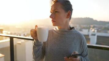 vrouw begint haar dag met een kop van thee of koffie en controle e-mails in haar smartphone Aan de balkon Bij ochtendgloren, langzaam beweging. modern stedelijk levensstijl video