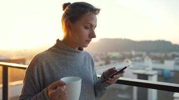 mujer empieza su día con un taza de té o café y comprobación correos electrónicos en su teléfono inteligente en el balcón a amanecer, lento movimiento. moderno urbano estilo de vida video