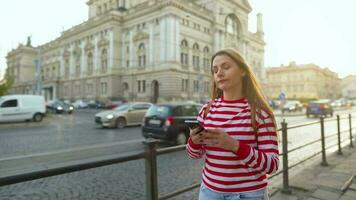 Jeune femme portant une rouge rayé chandail en marchant vers le bas un vieux rue en utilisant téléphone intelligent à le coucher du soleil. communication, social les réseaux, en ligne achats concept. video