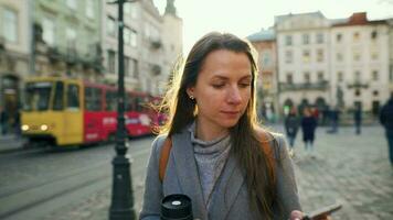 Woman wearing a gray coat with a thermos cup in hand walking down an old street using smartphone at sunset. Communication, social networks, online shopping concept. video