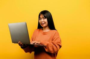 online communication concept asian girl use laptop at home yellow studio photo She is happy to spend her free time online while chatting with friends on social networks, shopping or working online.