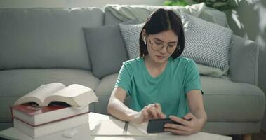 imágenes de cerca de joven asiático mujer lentes escritura en papel mientras acecho en teléfono inteligente a hogar. gente, e-learning y estilo de vida conceptos. video