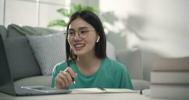 imágenes de cerca de joven asiático mujer vistiendo lentes escritura en papel mientras vídeo conferencia en el ordenador portátil cuaderno a hogar. gente, negocio y estilo de vida conceptos. video
