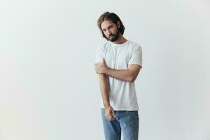 A man with a beard and long hair in a white T-shirt and blue jeans stands against a white wall leaning full-length on it, relaxed style photo