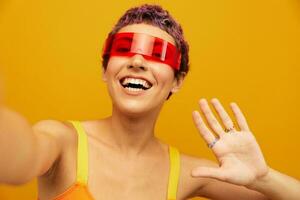 Woman wearing unusual millennial glasses taking selfies in sportswear against an orange studio background, free space photo