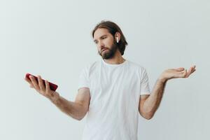 un hombre con un barba blogger en un blanco camiseta con un teléfono y inalámbrico auriculares mira dentro el teléfono y se extiende su manos a el lado en un blanco antecedentes aislado Copiar espacio foto