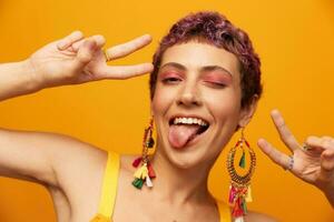 Portrait of a young woman with a short haircut and colored hair smiling and showing her tongue at the camera on an orange background with earrings accessories in the studio photo