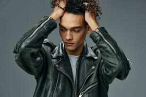 Portrait of stylish tanned curly man leather jacket touches head posing isolated on over gray studio background. Cool fashion offer. Huge Seasonal Sale New Collection concept. Copy space for ad photo