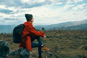 Traveler In a red jacket, hats with a backpack sits on a stone in the mountains in nature photo
