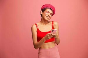Young woman blogger with colored pink hair and short haircut flipping through the phone screen and typing a message with a smile and surprise in stylish clothes on a pink background photo