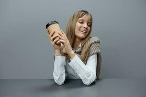 Cheerful cute young blonde student lady freelancer in warm sweater with takeaway coffee smiling looks aside in gray modern home office. Coffee break Lover Concept. Copy space. Cool offer photo