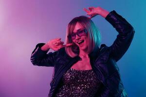 Happy beautiful blonde lady in leather jacket sparkly dress trendy sunglasses dance hold hands up posing isolated in blue pink color light studio background. Neon party Cyberpunk concept. Copy space photo