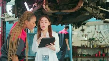 Young Asian female auto business entrepreneur discusses with mechanical worker, checklist undercarriage of lifted electric car for repair at maintenance garage, automotive service works industry. video