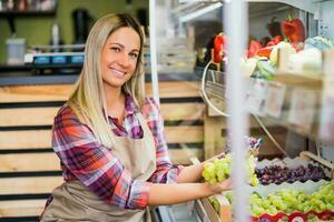 Woman works in fruits and vegetables shop. She is holding a cluster of grapes. photo