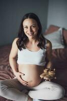 Pregnant woman relaxing at home. She is sitting on bed and holding baby shoes. photo