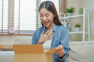 Happy excited, asian young woman, girl customer sitting on sofa at home, opening and unpacking cardboard box carton parcel after buying ordering present, shopping online, delivery service concept. photo