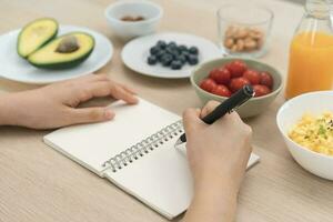 dieta, dieta mano de asiático joven mujer, niña escribir dieta plan nutrición en mesa es diferente comida ingredientes en el verde. nutricionista de saludable, nutrición de peso pérdida, salud cuidado gente. foto