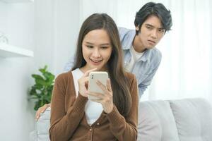 Infidelity, suspicion asian young couple love, wife using mobile phone, husband watching spying his girlfriend while woman chat a message, man sneaky distrust and jealousy, relationship problem. photo