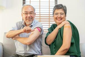 Coronavirus Vaccination, happy asian elderly, aged family smile strong together, showing bandage on arm with protect of covid-19 after injection of vaccine, sitting on couch in living room at home. photo