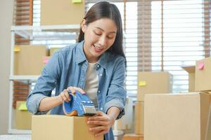 Small business entrepreneur SME, asian young woman,girl owner packing product, checking parcel for delivery to customer, using scotch tape to seal the box, working at home. Merchant online, e commerce photo