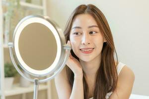 Fresh healthy skin, beautiful smile of asian young woman, girl looking at mirror, applying moisturizer on her face, putting cream treatment before make up cosmetic routine at home. Facial Beauty. photo