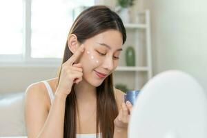 Fresh healthy skin, beautiful smile of asian young woman, girl looking at mirror, applying moisturizer on her face, putting cream treatment before makeup cosmetic routine at home. Facial Beauty. photo