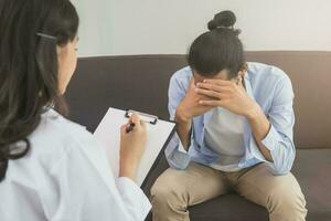 Asian young man, male has mental symptoms which must have been therapy and stress, sitting on couch to consult to psychologist during the session  taking notes to find out how to treat the therapist. photo