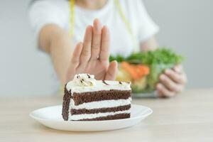 dieta, dieta asiático joven mujer utilizando mano empujar afuera, negar chocolate pastel o dulce gusto, postre alimento, escoger comer verde ensalada vegetales cuando hambriento. nutricionista de saludable, nutrición de peso pérdida. foto