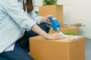 Focus hand of asian woman, female holding packing, unpacking box by tape machine, sealing cardboard indoors. Business owner in warehouse, Moving or preparing in new house, apartment or relocation. photo