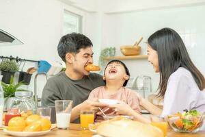 contento refresco familia desayuno en mañana, asiático joven padre padre, madre y pequeño linda chico, niño teniendo comida en cocina comiendo juntos a hogar. alegre, disfrutar Cocinando gente. foto