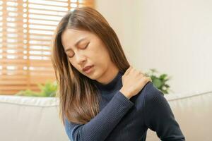 Body muscles stiff problem, asian young attractive woman, girl pain with back pain ache from work, holding massaging rubbing shoulder hurt or sore, painful sitting on sofa at home. Healthcare people. photo