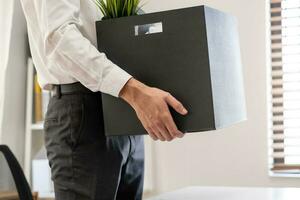 Unemployed, quite job. Desperate caucasian young businessman resigning from company, hand in holding cardboard, packing belongings into box, layoff or changing work. Resignation, employment concept. photo