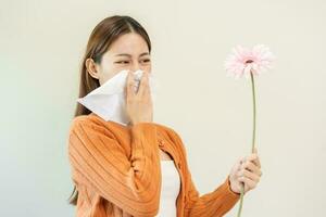 Allergic rhinitis symptom of odor pollen flowering, portrait asian young woman hand in sneeze, blowing runny nose after smell, smelly from bloom flower, holding away pink flora on white background. photo