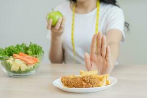 dieta, dieta asiático joven mujer mano en empujar afuera, negar frito pollo, francés frito y escoger verde manzana, vegetales ensalada, comer comida para bueno saludable, salud cuando hambriento. hembra peso pérdida gente. foto