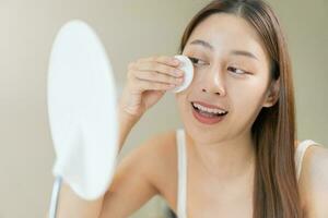 contento belleza, hermosa asiático joven mujer, niña mirando en a espejo, participación algodón almohadilla, aplicando facial limpiar en su rostro, quitando maquillaje antes de ducha en baño, piel cuidado en blanco antecedentes. foto