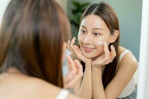 Fresco sano piel, hermosa sonrisa de asiático joven mujer, niña mirando a espejo, aplicando hidratante en su rostro, poniendo crema tratamiento antes de maquillaje cosmético rutina a hogar. facial belleza. foto