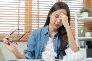 Exhausted, stressed asian young business woman working, took off glasses, massage nose from dry eyes, suffer on hard work while use laptop computer at office home. Overtime job, debt problem people. photo