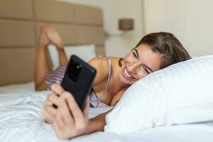 Smiling woman looking at mobile phone while laying on white bed. Happy brunette young woman using cellphone at home. Beautiful girl surfing the net on smartphone photo