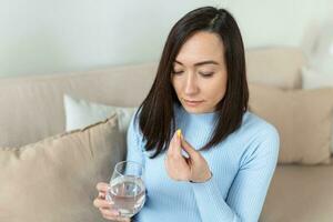Asian woman takes pill with glass of water in hand. Stressed female drinking sedated antidepressant meds. Woman feels depressed, taking drugs. Medicines at work photo
