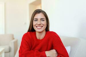 Happy young woman sitting on sofa at home and looking at camera. Portrait of comfortable woman in winter clothes relaxing on armchair. Portrait of beautiful woman smiling and relaxing during autumn. photo