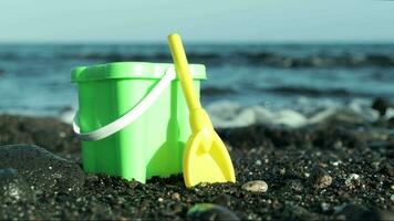 closeup of a plastic bucket and spade on a black sand beach with the ocean in the back ground video