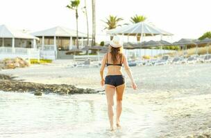 mujer en un sombrero y traje de baño caminando en el playa en un caliente verano día - ver desde el espalda foto