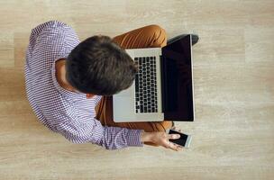 Top view of man sitting on the floor and working with a laptop and smartphone in his hand photo