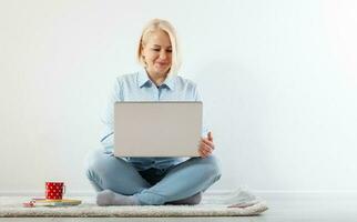 Happy middle aged woman sitting relaxed on the floor using laptop for entertainment. The concept of leisure and work with a cup of coffee at home. photo