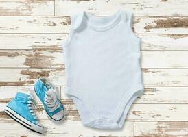 Mockup of white baby bodysuit on white wood background with sneakers. Blank baby clothes template in farmhouse style, flat lay. photo