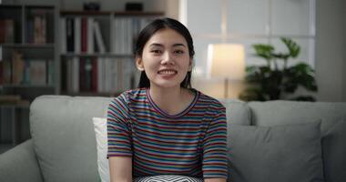 Close up shot, front view, Asian young pretty woman sitting on comfortable sofa and watching television, she smile while sing a song karaoke for relax in living room with happy at home video