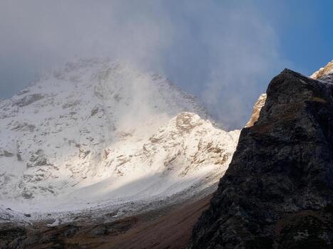 Atmospheric landscape with sharp rocks and high snowy mountain top in blizzard low clouds at overcast. Dramatic gloomy scenery with large snow mountains and glacier in gray cloudy sky at snowy weather photo